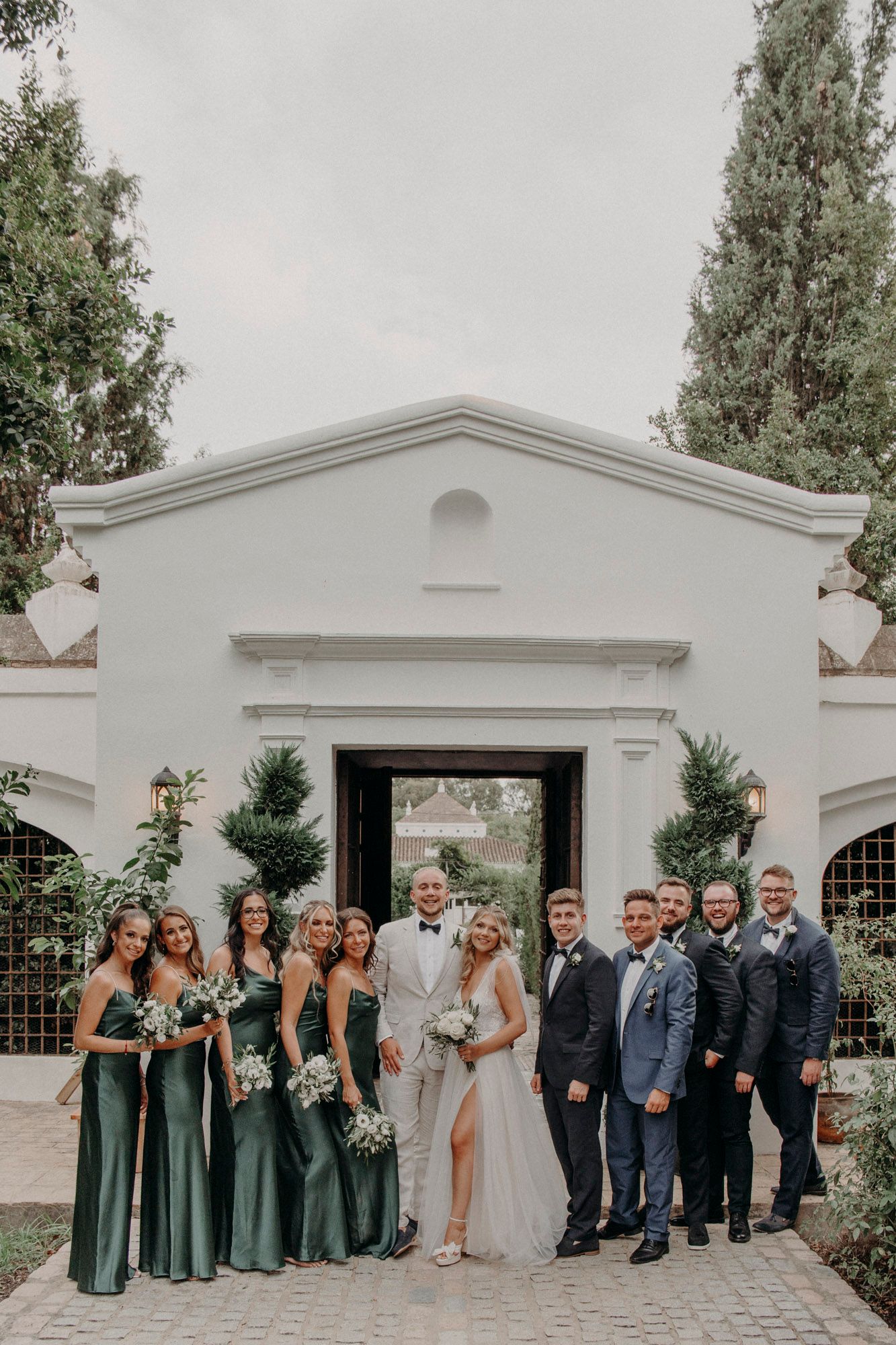 Wedding party in emerald green dresses and dark suits pose in front of elegant white building with arched entrance.