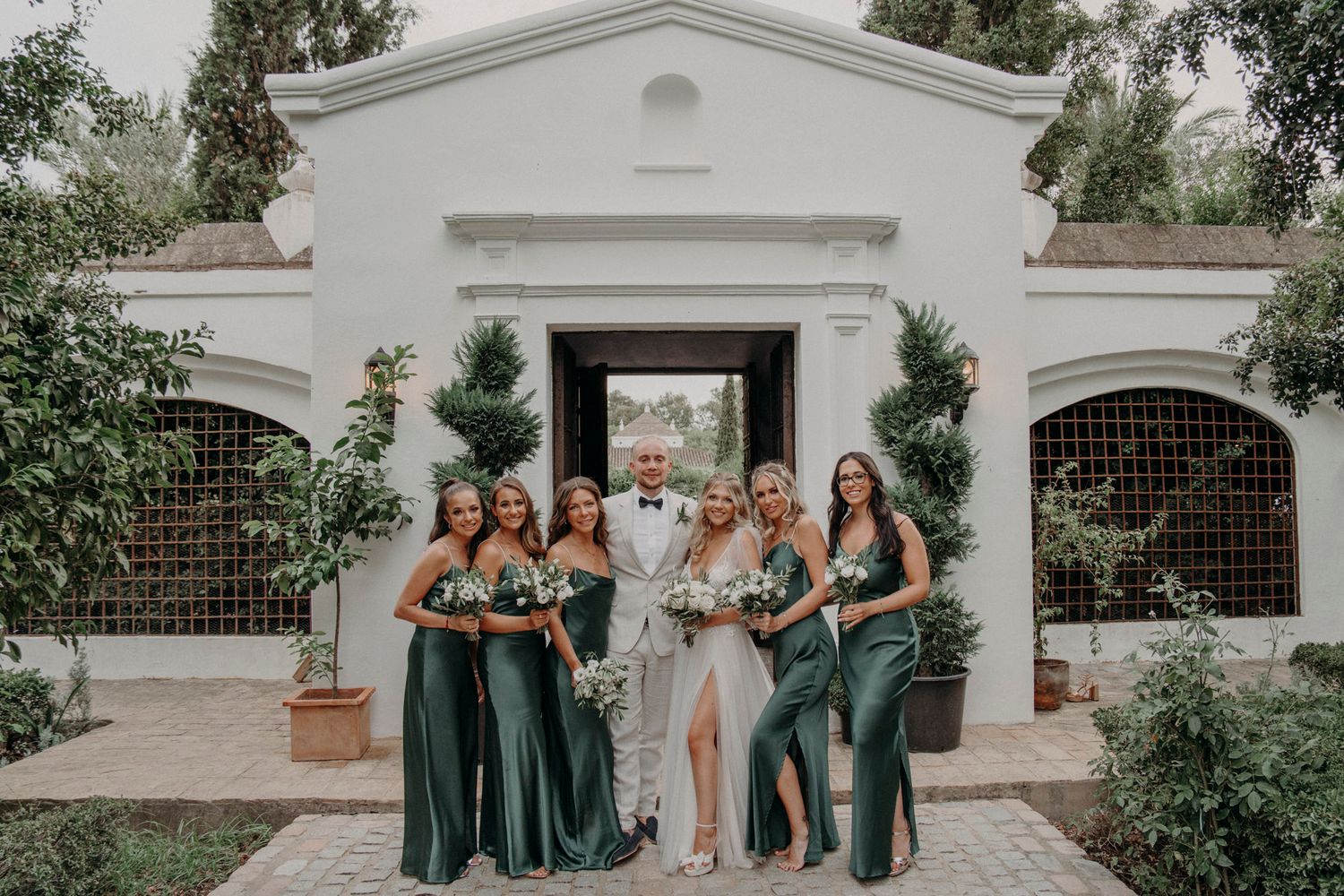 Wedding party in emerald green dresses poses together outside ornate white Spanish-style building entrance.