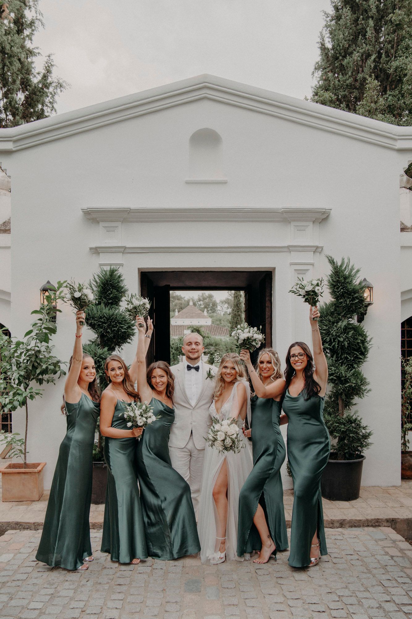 A wedding party poses in front of a white Mediterranean-style doorway with greenery and florals.