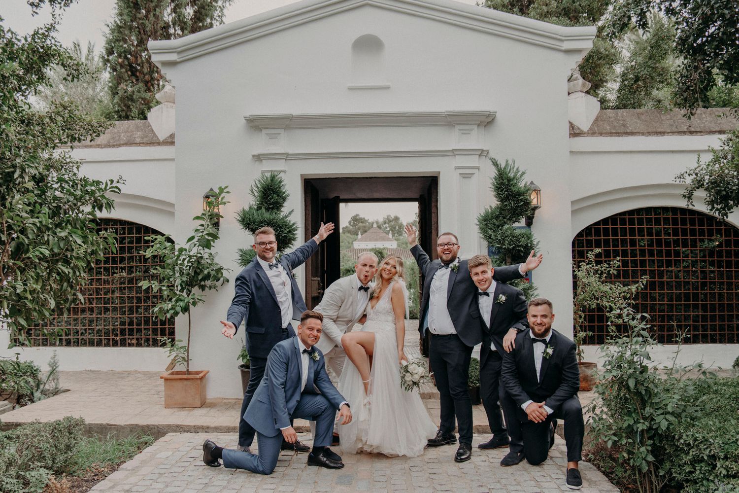 Wedding party poses playfully outside elegant white Spanish-style building with arched windows and greenery.