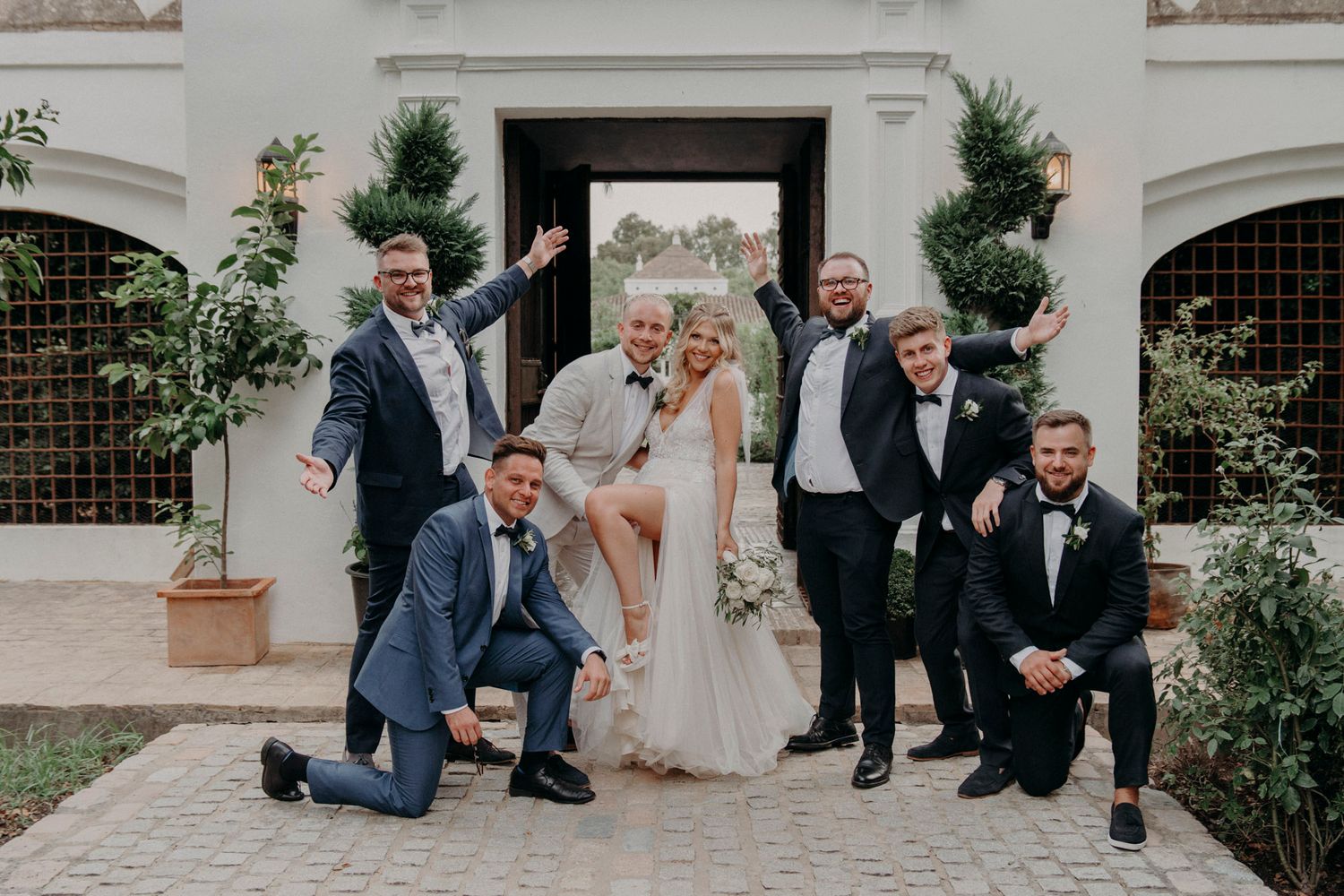 Wedding party poses playfully outside a white building entrance with decorative topiary plants.