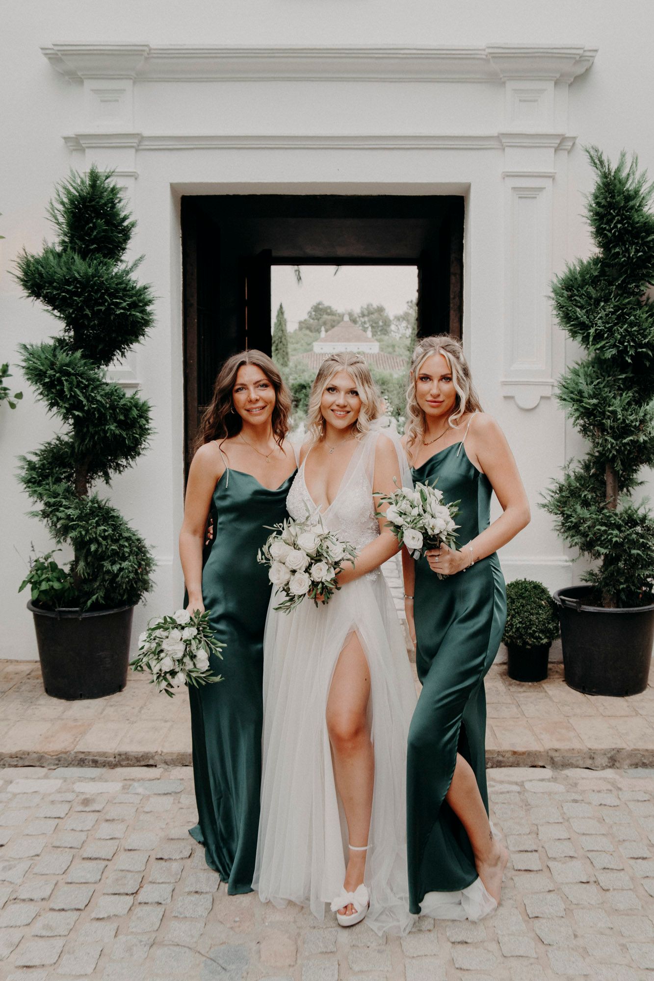 Bride in white gown poses with bridesmaids wearing emerald green silk dresses by elegant doorway.