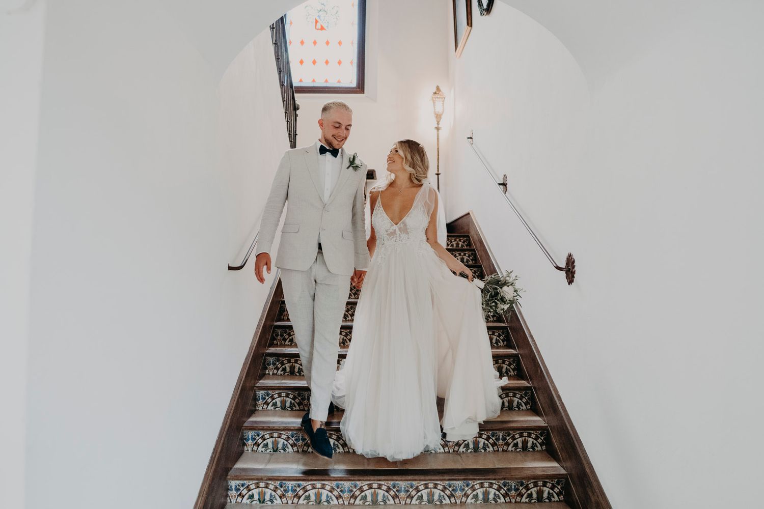 Wedding couple in white attire descends ornate staircase with decorative railing in elegant venue.