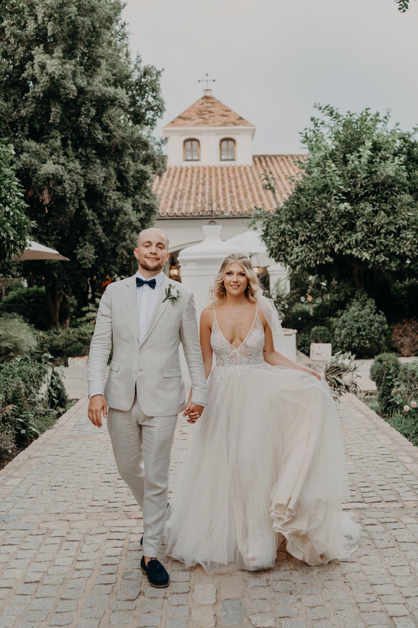 A couple walks hand in hand down a cobblestone path with a Spanish mission style church in the background.
