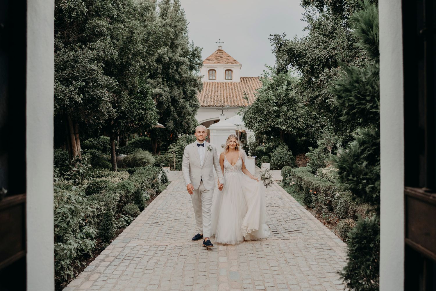 Newlyweds walk down garden path with Spanish-style church building in background.