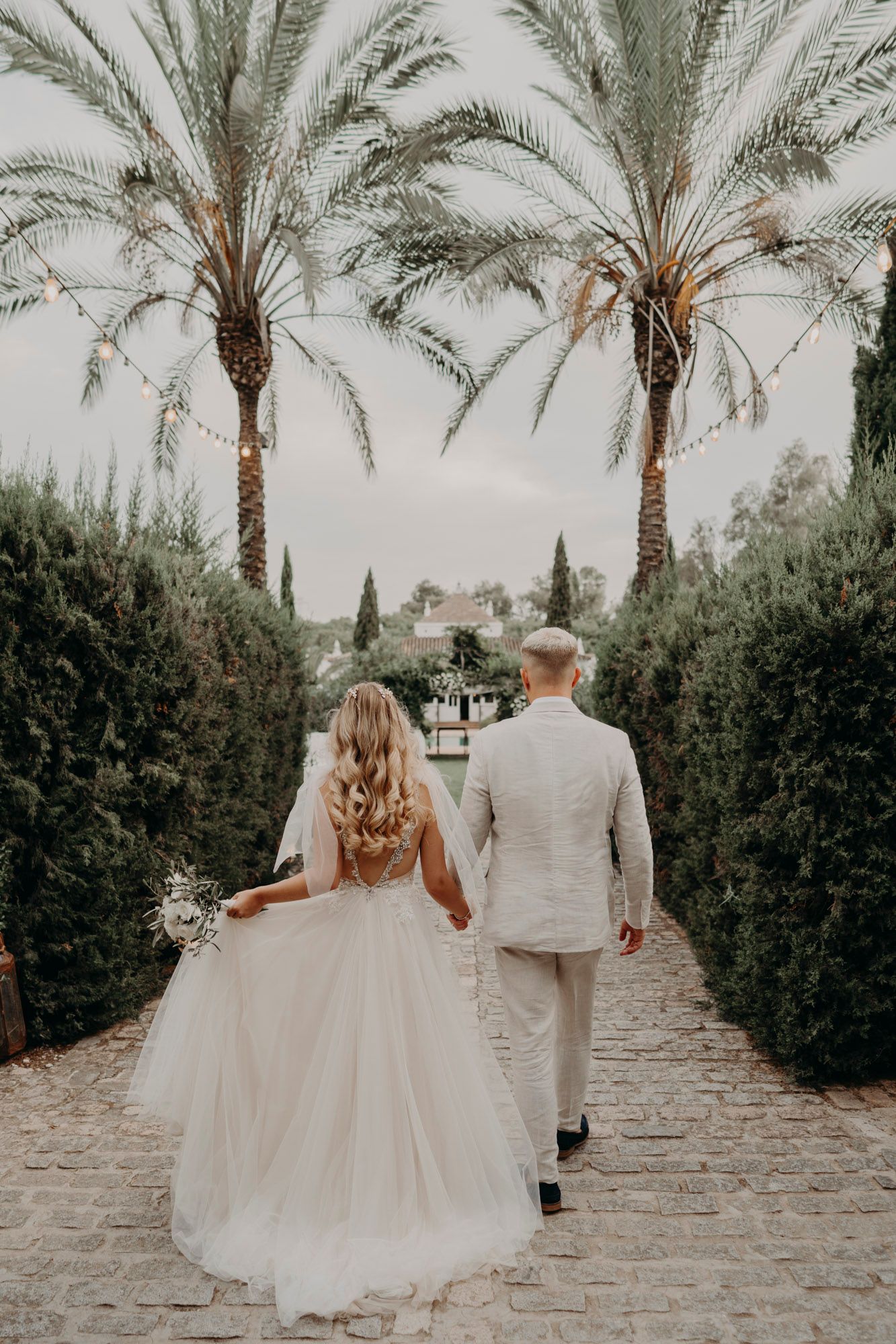 A wedding couple walks down a palm tree-lined stone pathway holding bouquets in a Mediterranean garden setting.