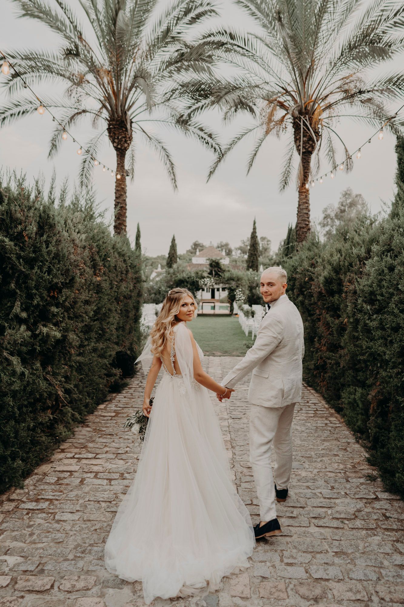 Wedding couple walk hand in hand down palm-lined stone pathway in romantic garden setting.