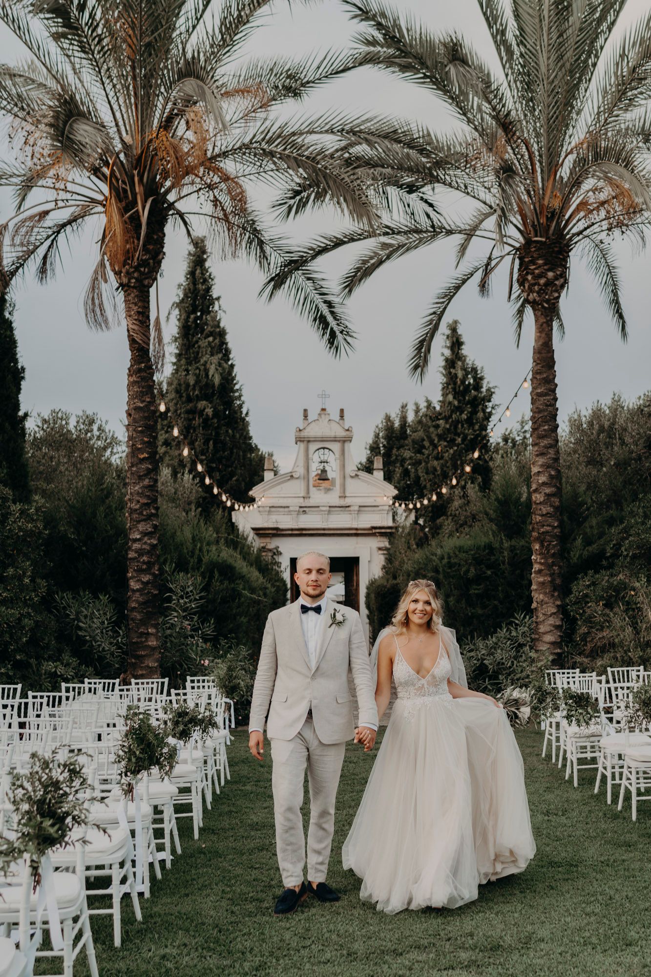 Elegant outdoor wedding ceremony setup with palm trees framing white chairs and a classical architectural backdrop.