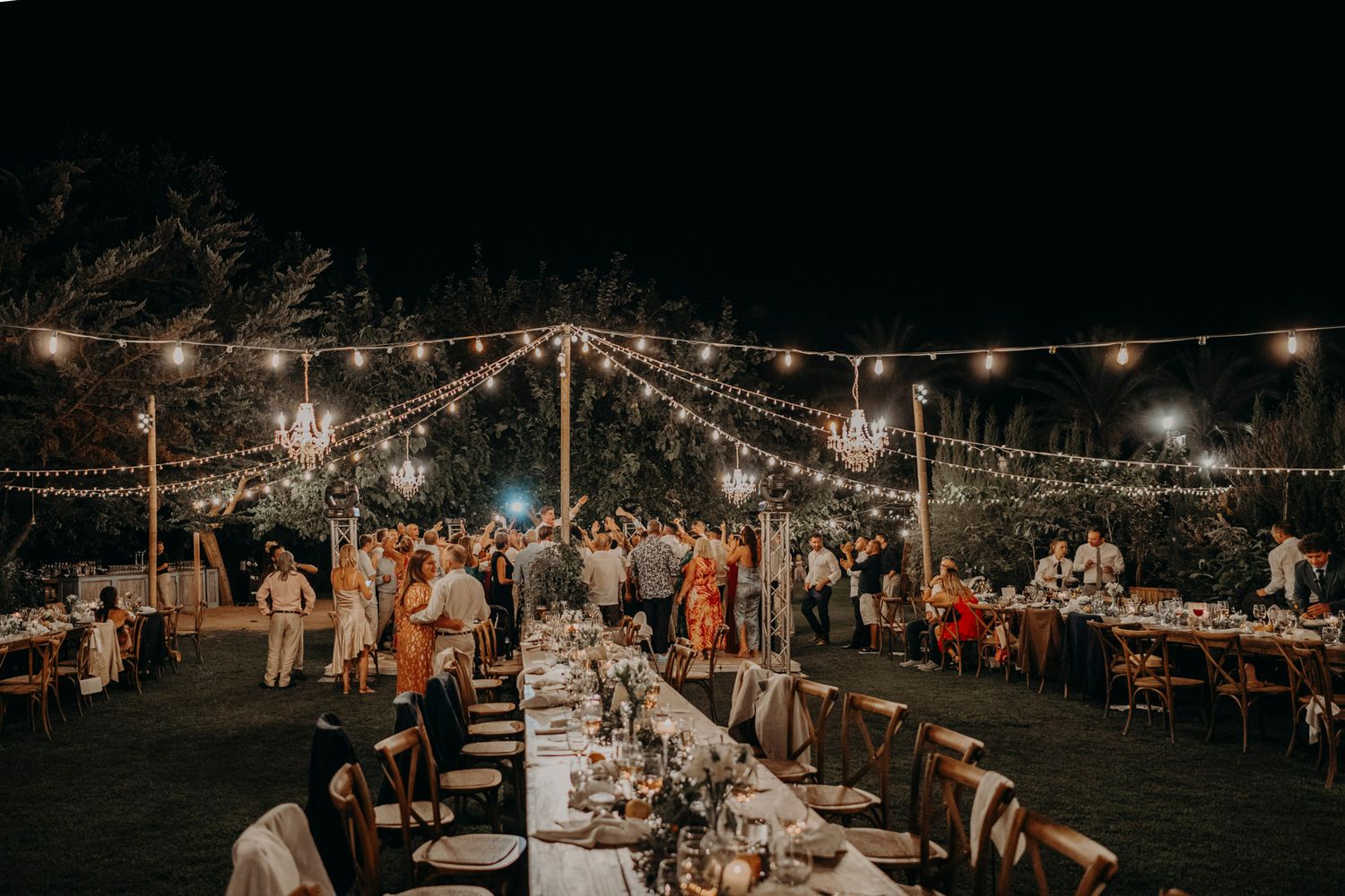 An outdoor evening wedding reception under string lights with long dining tables and guests mingling in a garden setting.