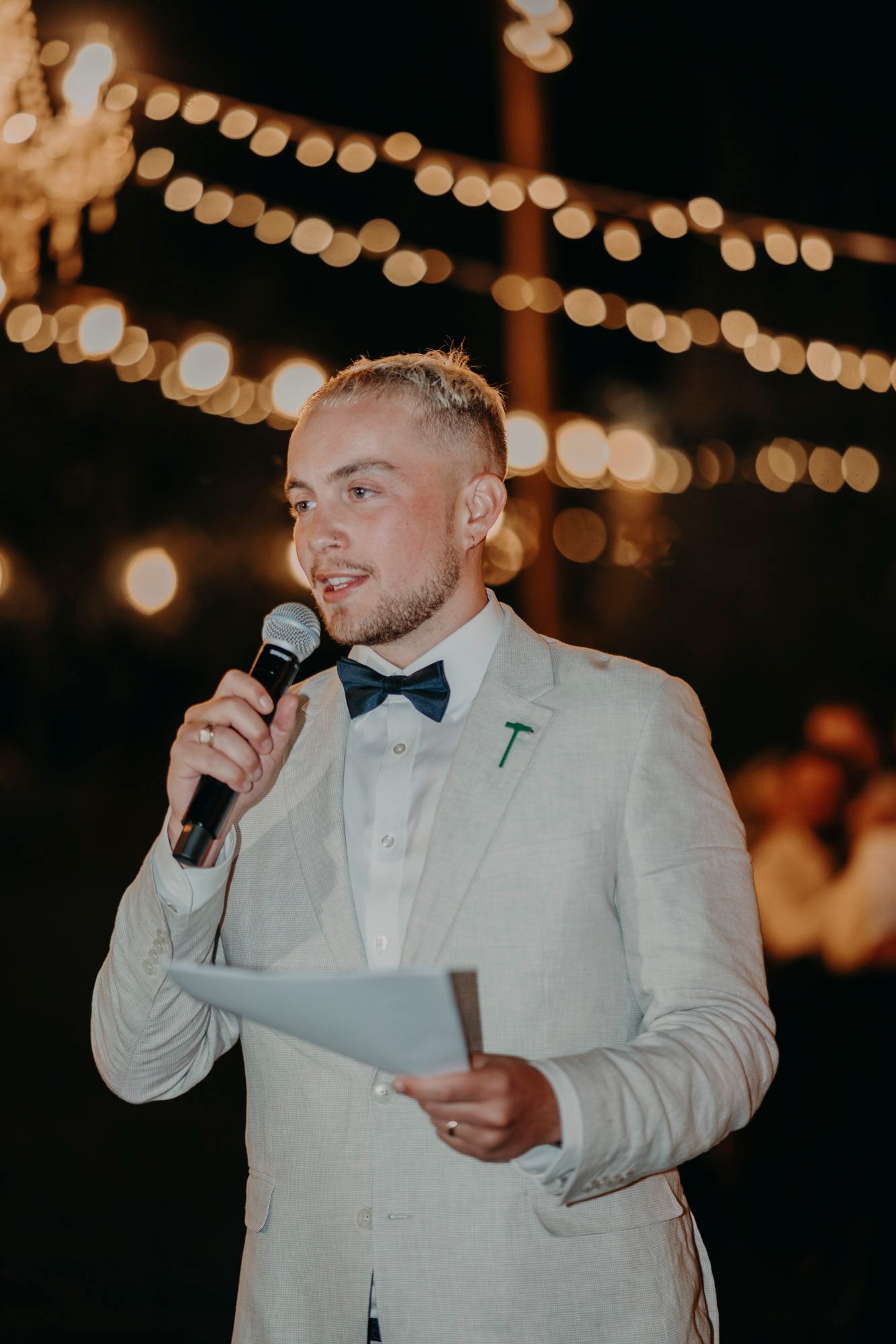 Wedding guest gives speech at outdoor evening reception under glowing string lights, wearing light colored suit and bowtie.