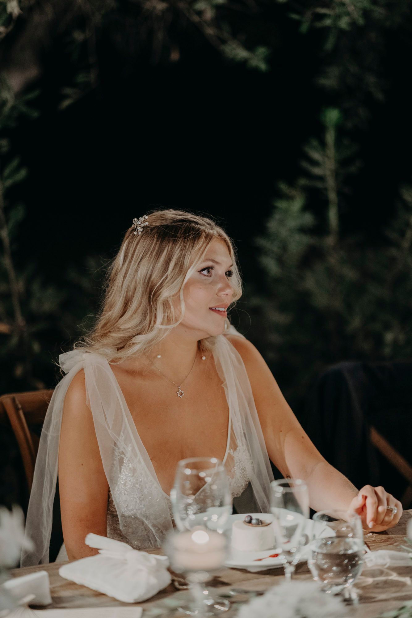 Elegant bride in white dress sits at reception table glancing to the side with natural backdrop.