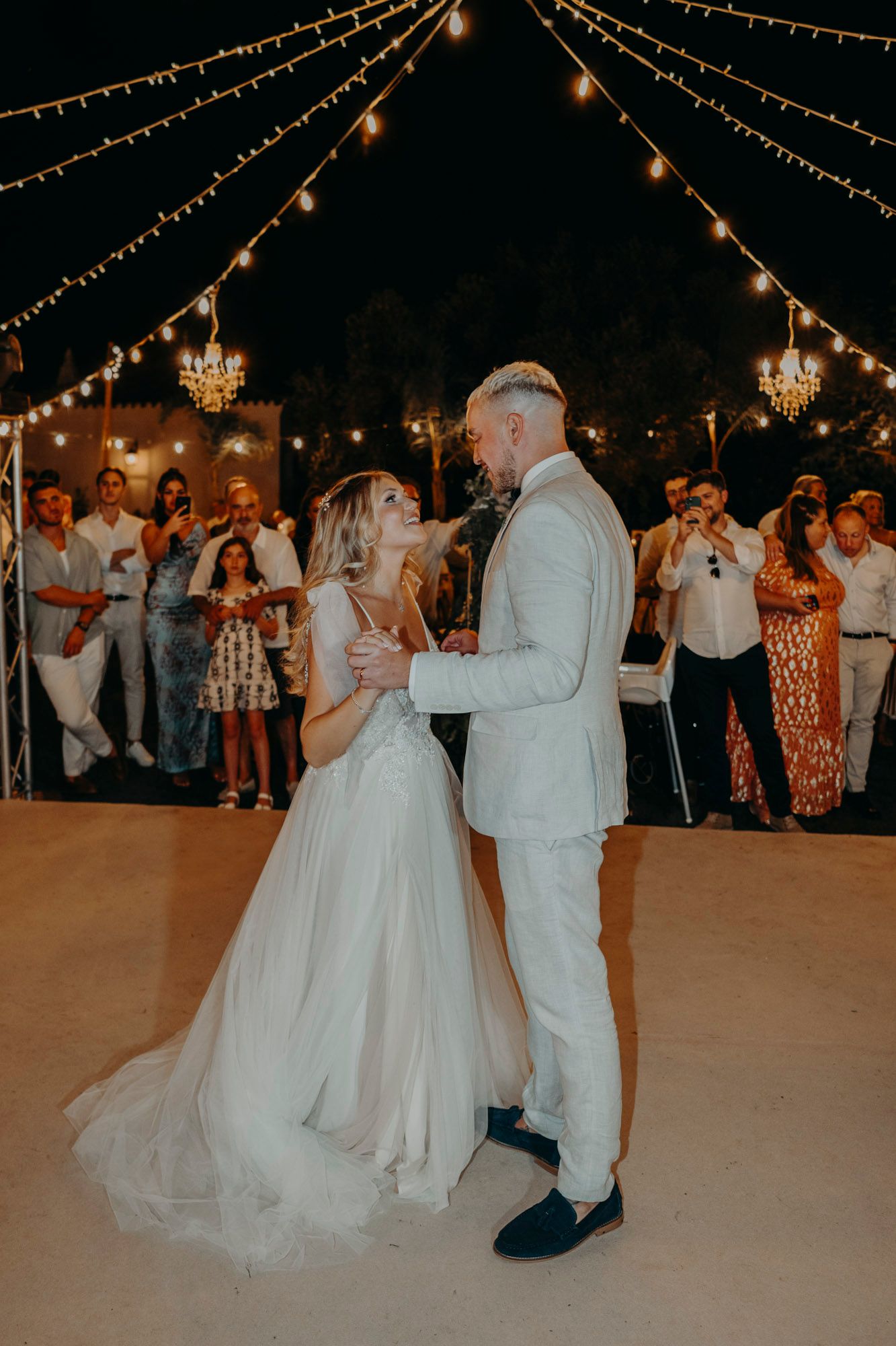 A couple shares their first dance under romantic string lights at an outdoor evening wedding reception.