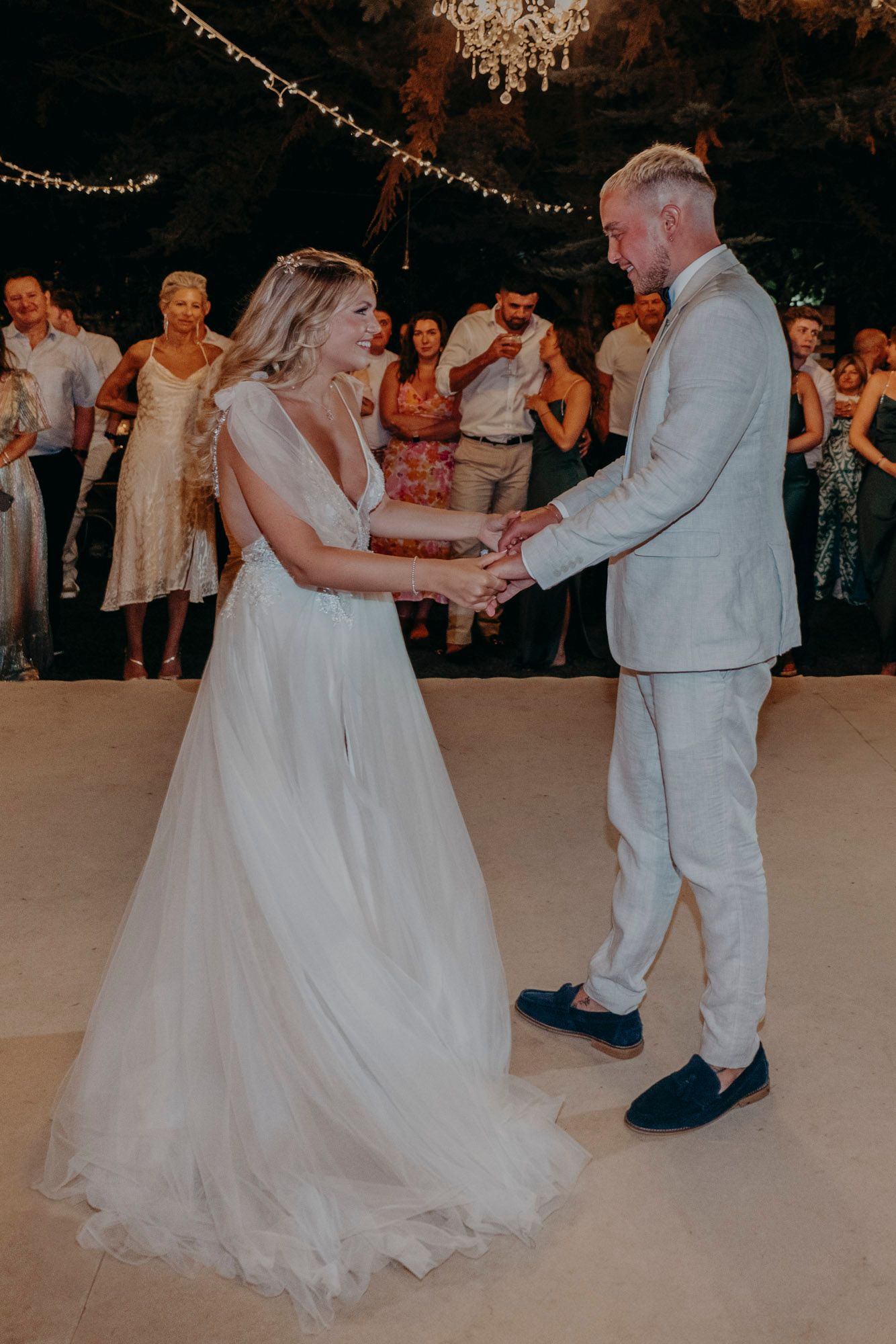 A bride and groom share their first dance under twinkling lights at an indoor wedding reception with guests watching.