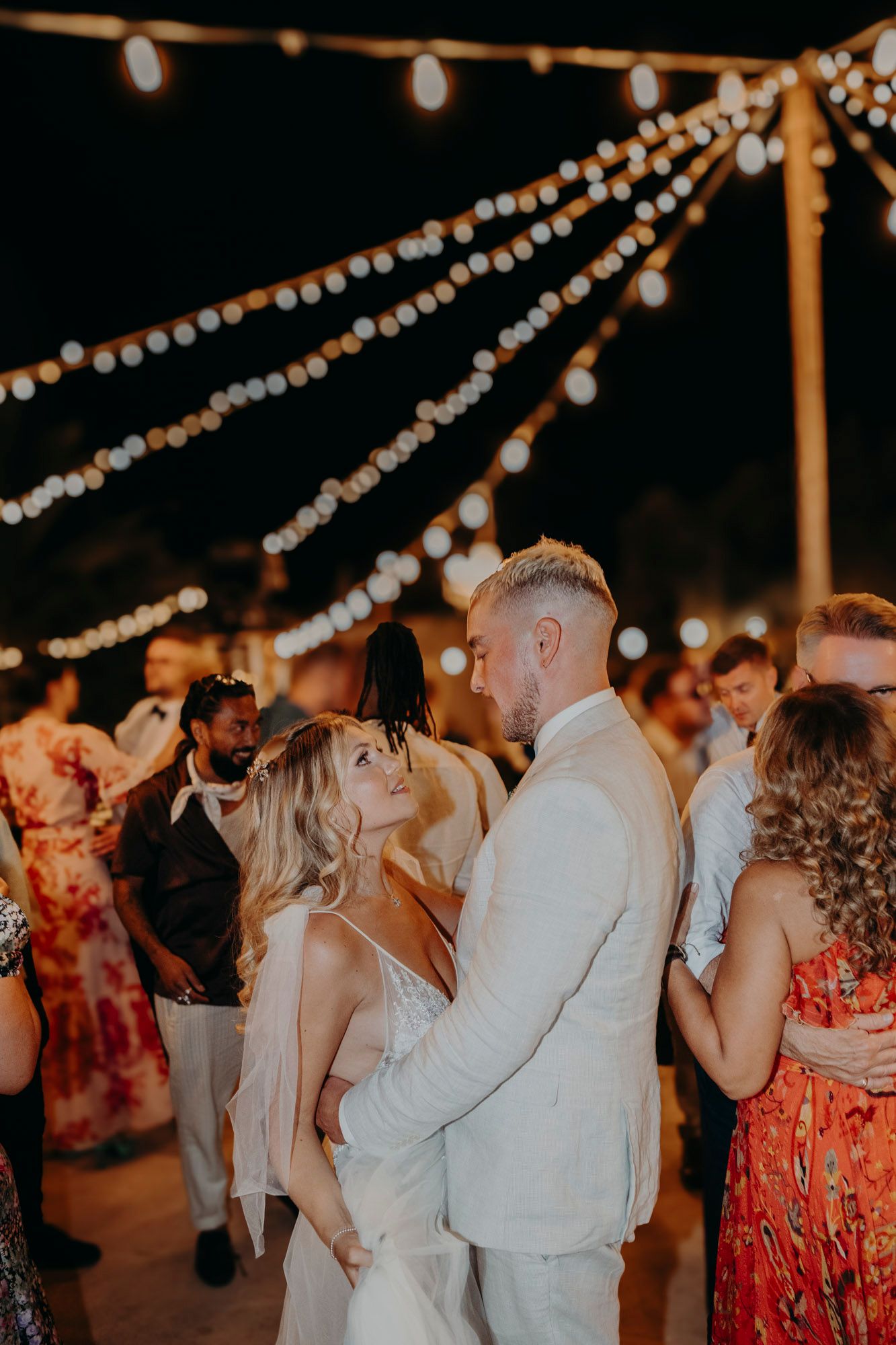 Couples dance under romantic string lights at an outdoor wedding reception with festive floral dresses and summer attire.
