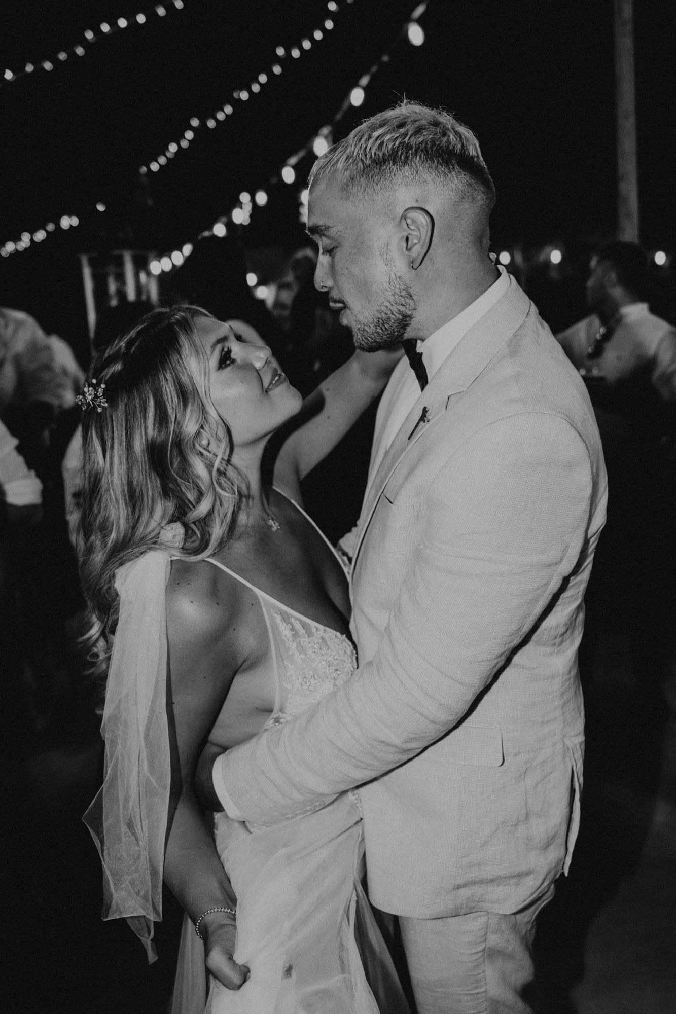 A romantic black and white photo of a couple in wedding attire sharing an intimate moment under string lights at night.