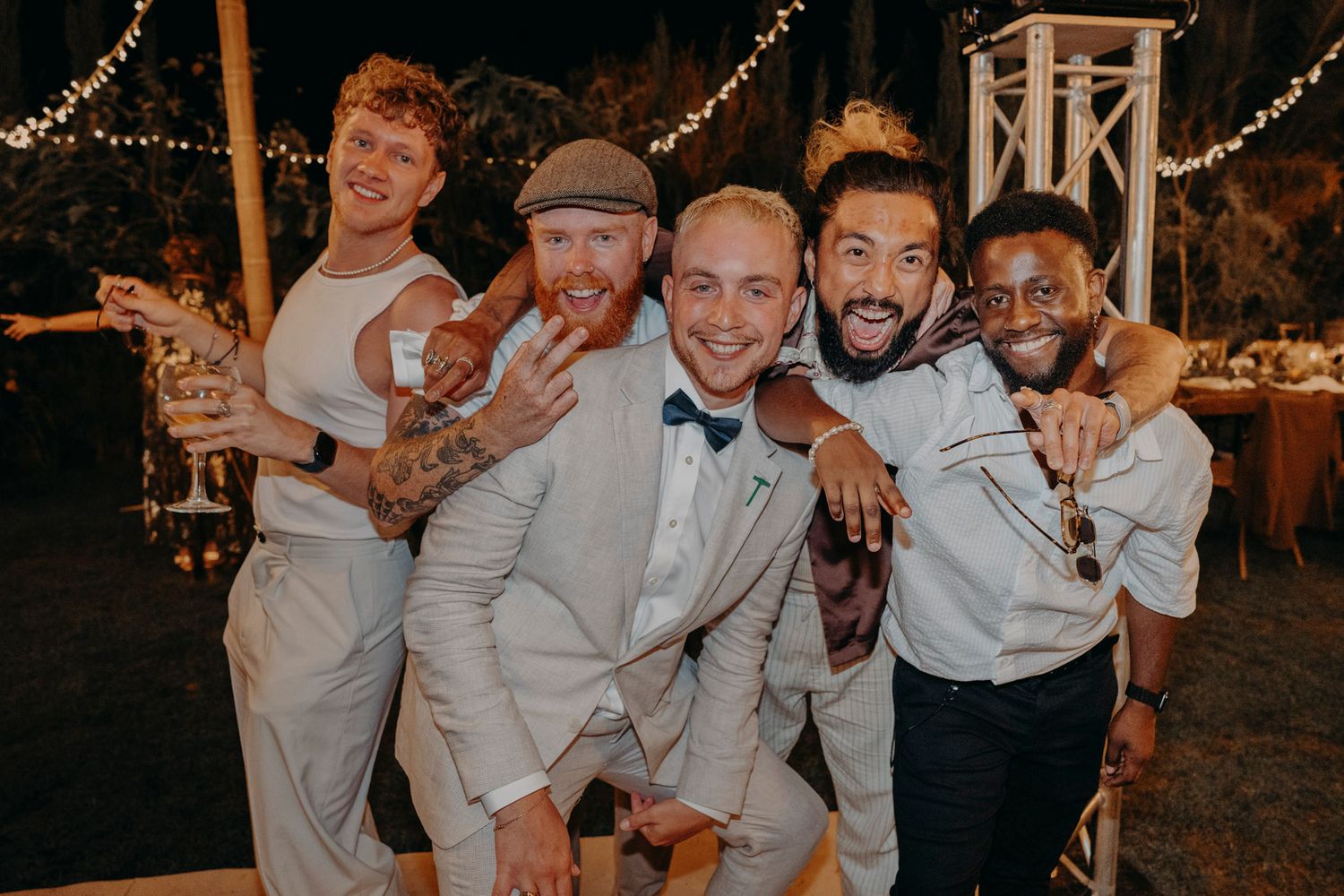 Group of friends in formal attire pose together at an outdoor evening wedding reception with string lights glowing behind them.