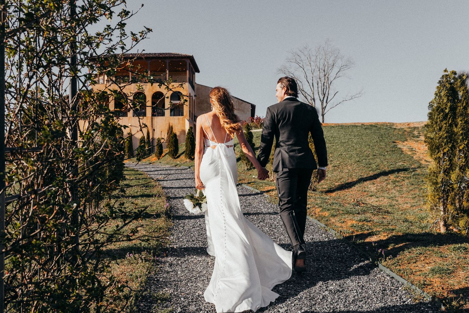 Candid wedding photo of couple walking along path toward rustic Italian villa.