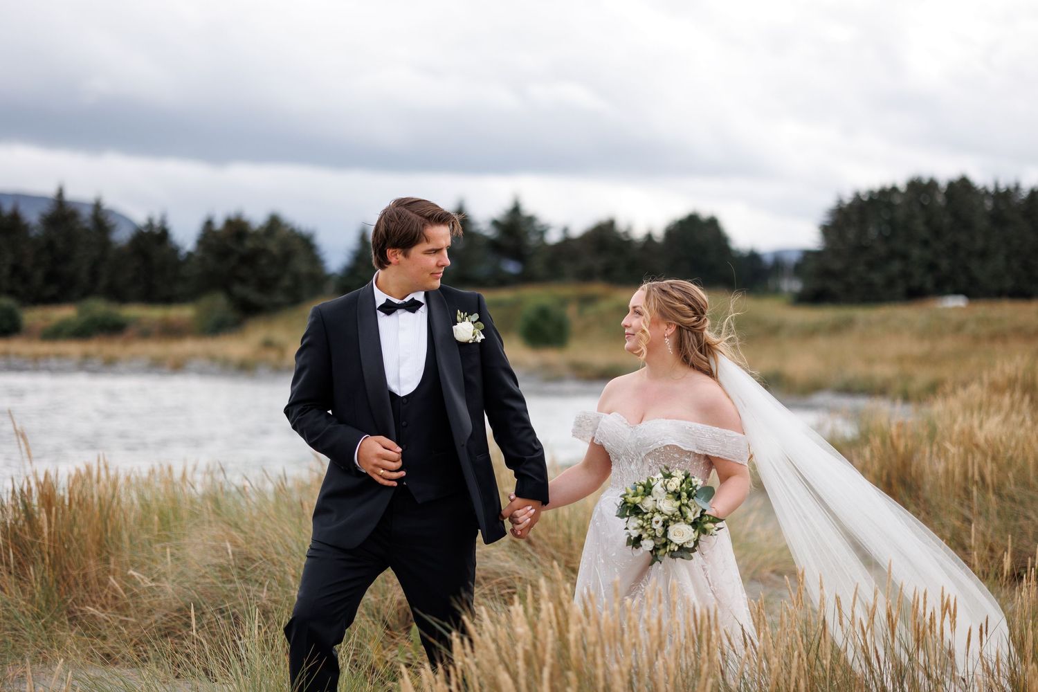 Newlywed couple walks through tall grass field near a lake in formal wedding attire.