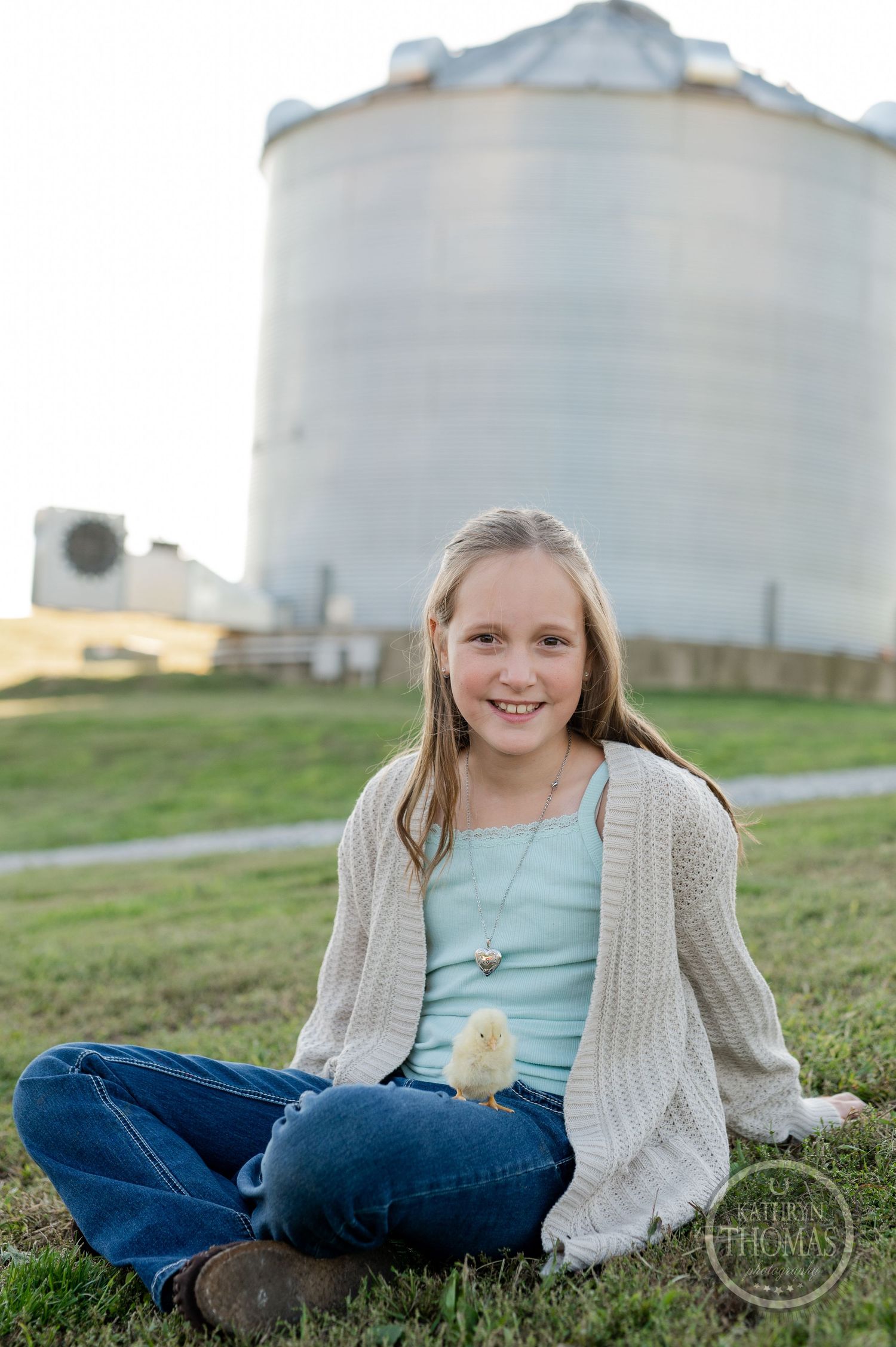 A person in a cream cardigan sits cross-legged on grass with a large grain silo in the background.