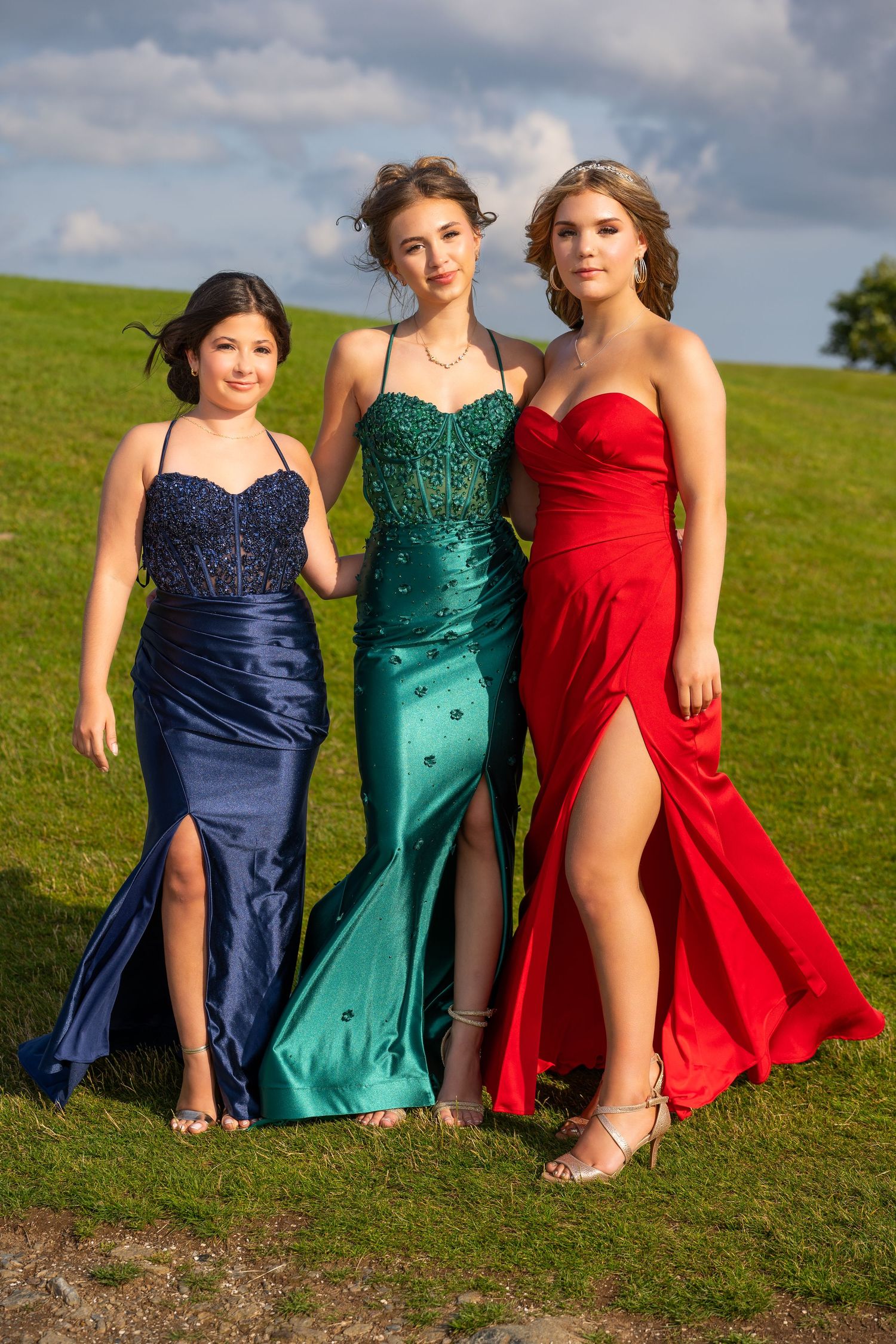 Three women pose together wearing elegant navy, emerald and red formal dresses on grassy hillside.