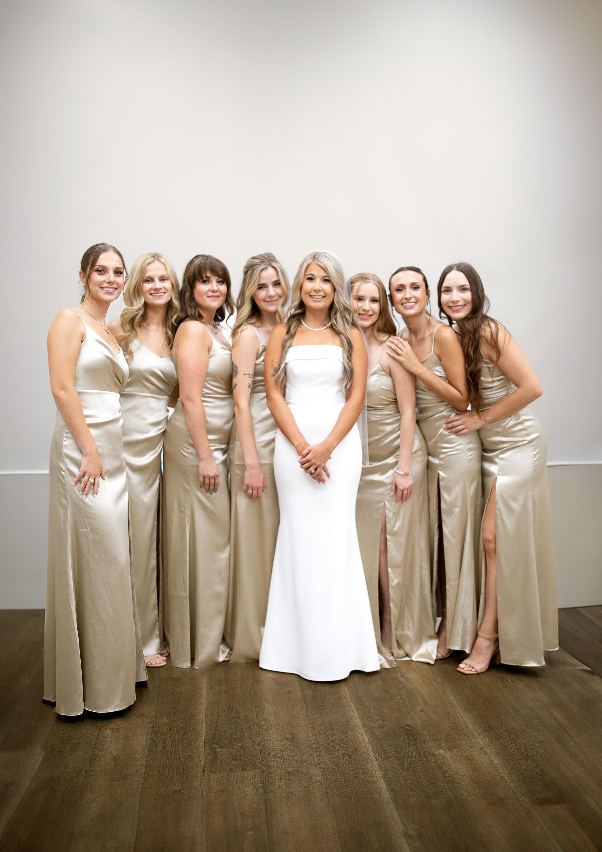 Bridal party in champagne satin dresses poses together against white wall.