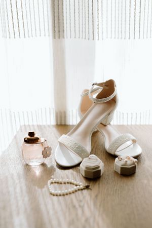 Wedding accessories including white heels, perfume bottle, and jewelry arranged on wooden surface.