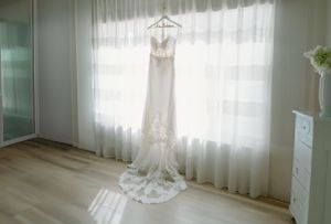 Elegant lace wedding dress hanging by window in bright, minimal room.