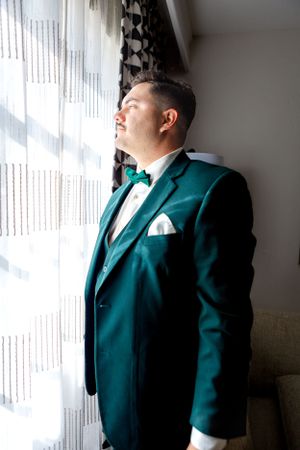 Elegant groom in emerald green suit jacket looking out window.
