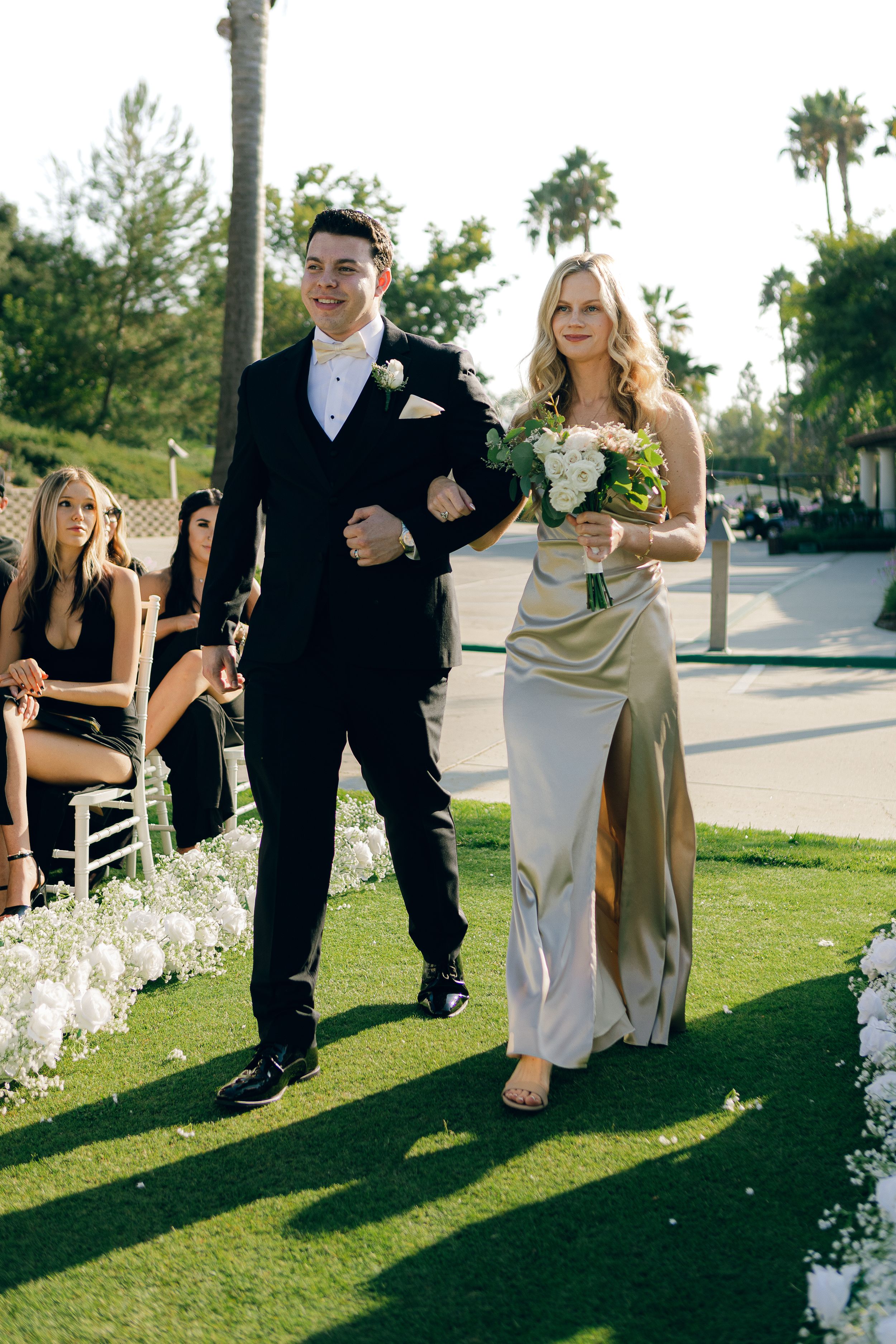 Wedding party members walk down outdoor aisle decorated with flower petals and white chairs.