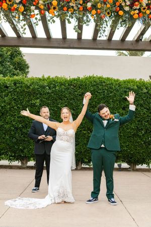 Wedding celebration under a floral pergola decorated with vibrant orange and white flowers against green hedge backdrop.