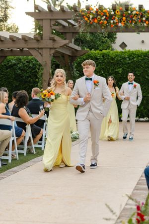 Wedding party members walk down aisle under wooden pergola adorned with bright floral arrangements.