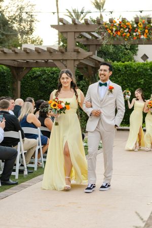 Couple walks down aisle after ceremony under pergola with yellow and orange floral decorations.