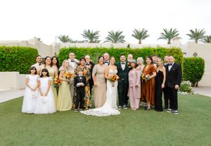 Large wedding party poses for group photo on grass lawn with palm trees and hedges in background.