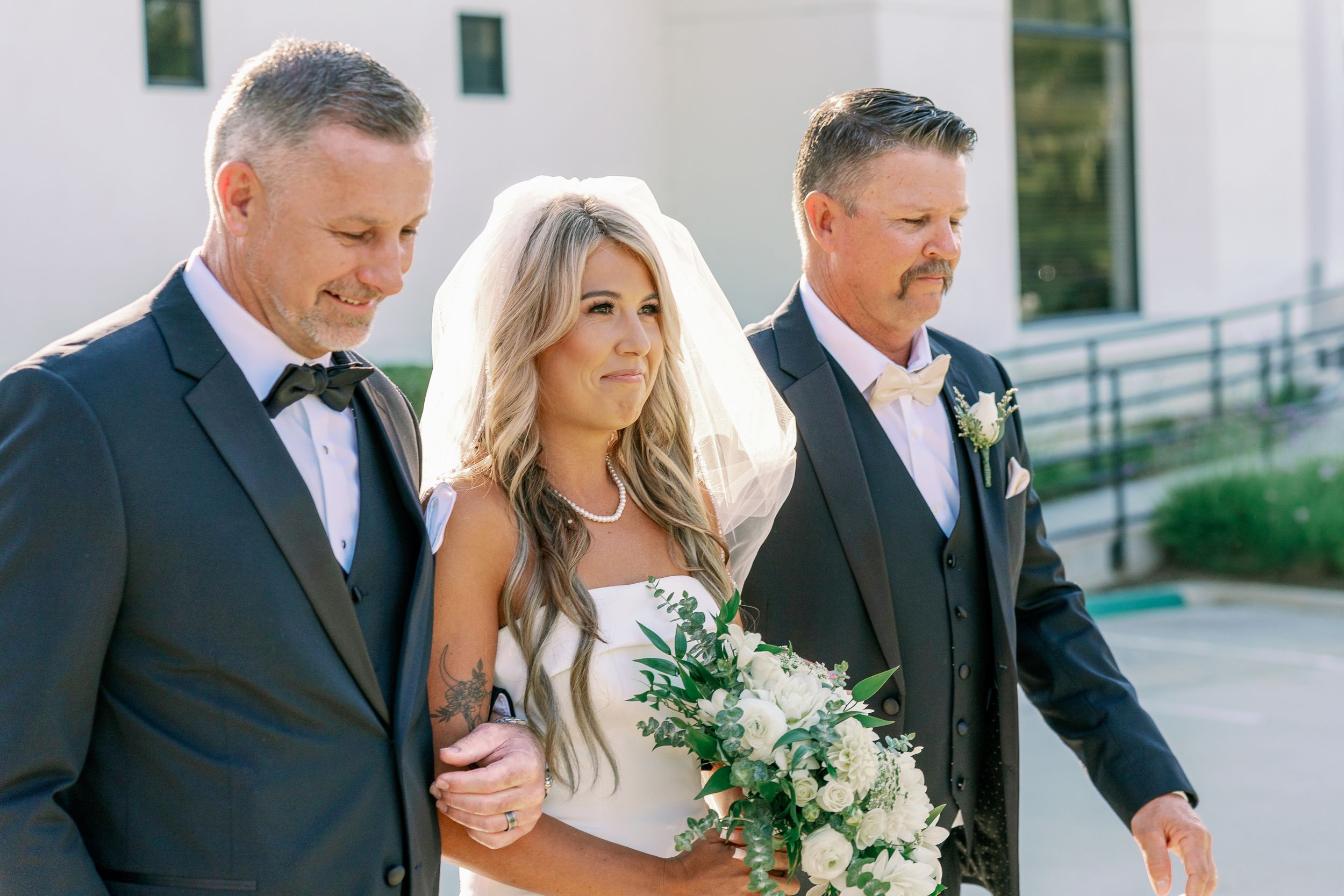 Groomsmen in black tuxedos pose together on leather bench in wood-paneled room.