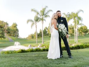 Newlyweds stand together on manicured golf course with fountain and palm trees in background.