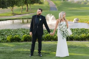 Elegant couple in wedding attire hold hands while walking along golf course with water features.