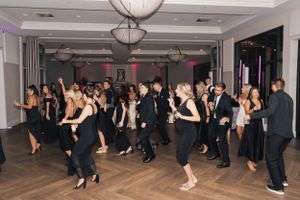 Large group of wedding guests dancing on wooden floor at reception under modern lighting fixtures.