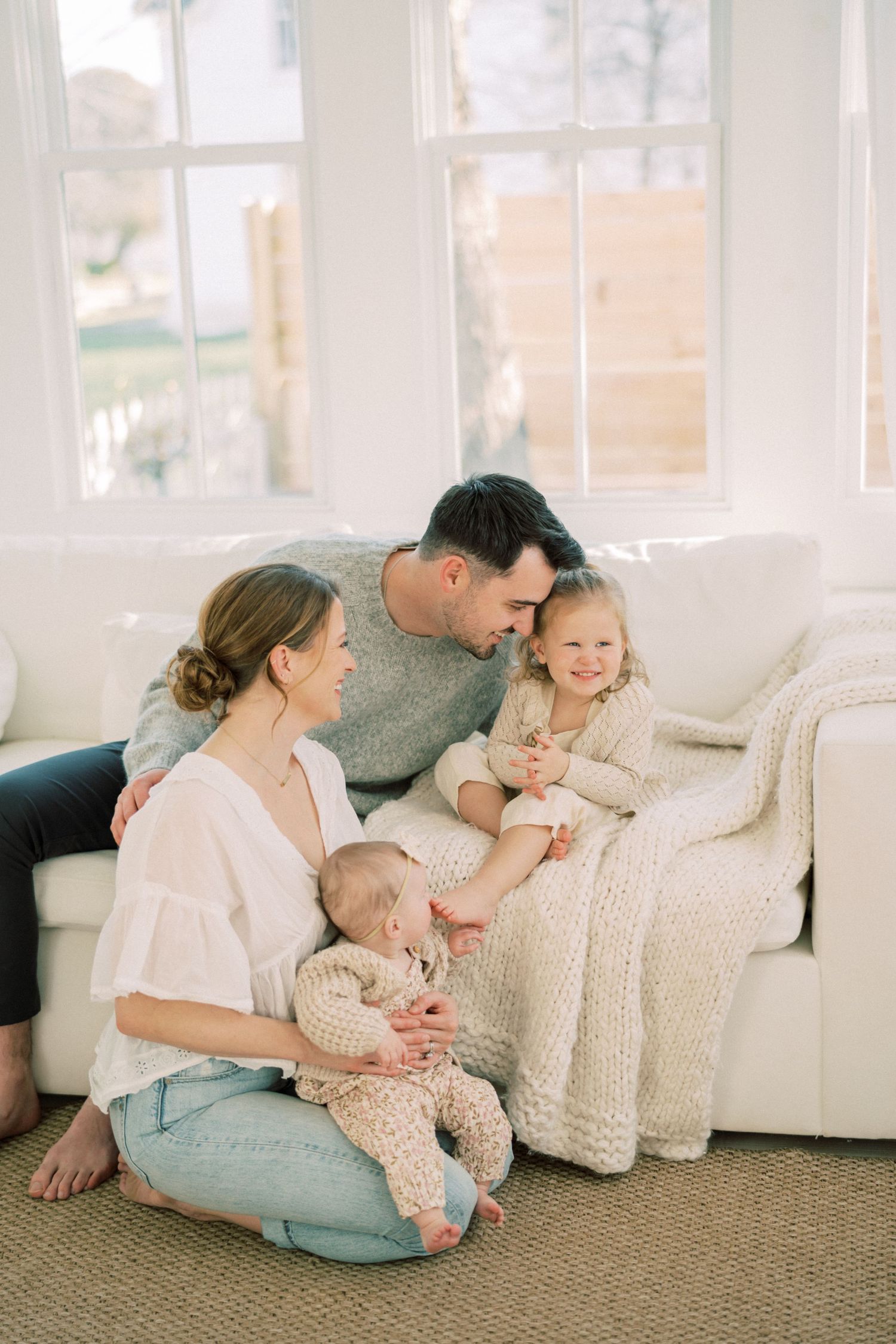 Cozy family moment on white sofa in bright sunlit room with toddler and baby in matching cream knit outfits.