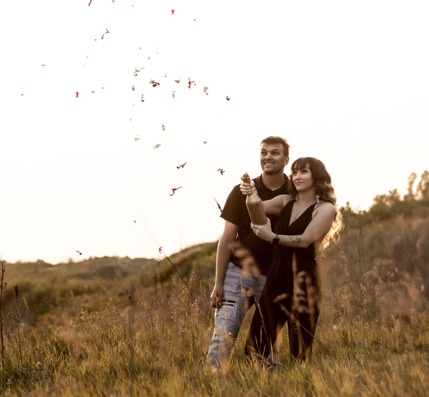Couple embracing in a field with confetti falling around them at sunset.