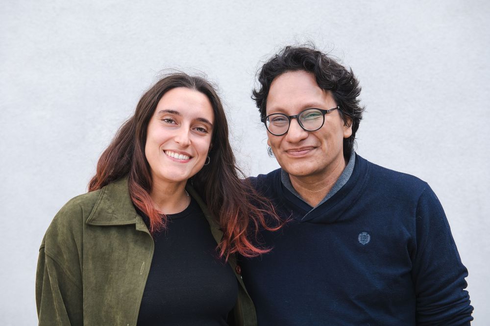 Two people in casual attire smile together against a light background.
