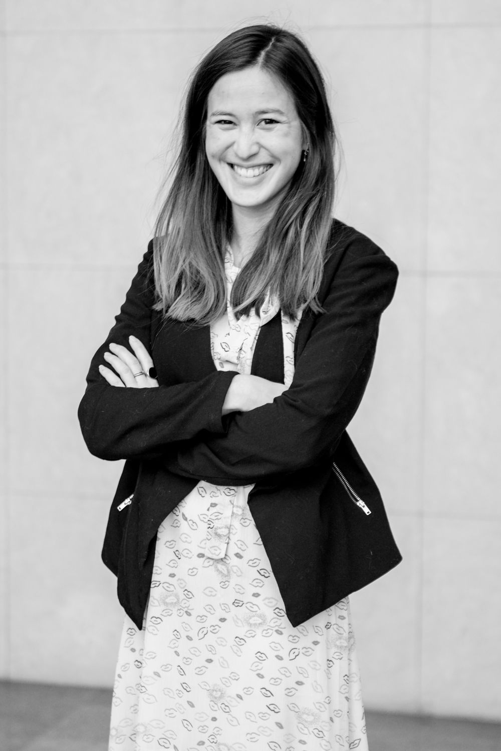 Person in black blazer and patterned dress poses playfully against light wall.
