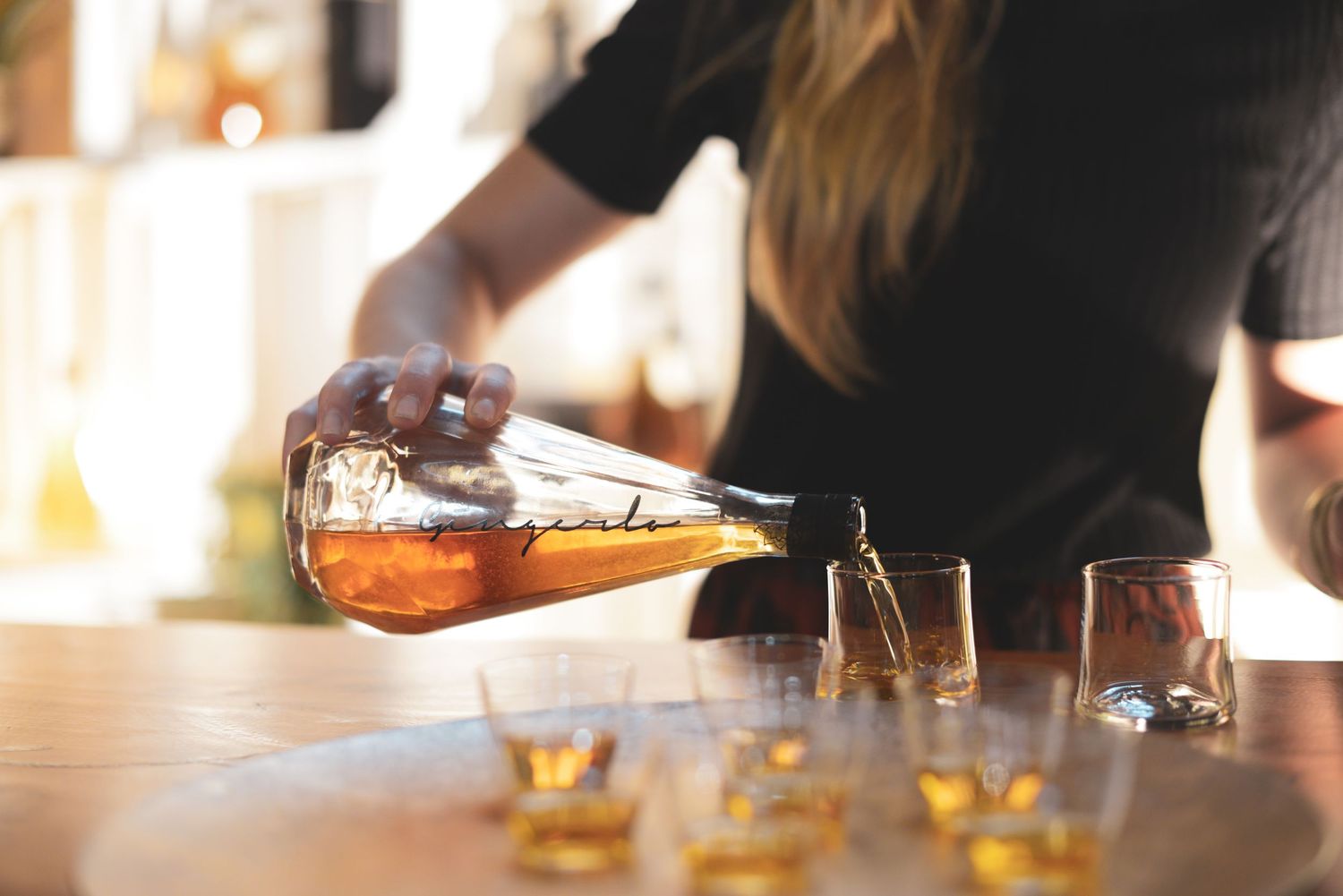 Row of whiskey glasses on a wooden bar counter with people in background.