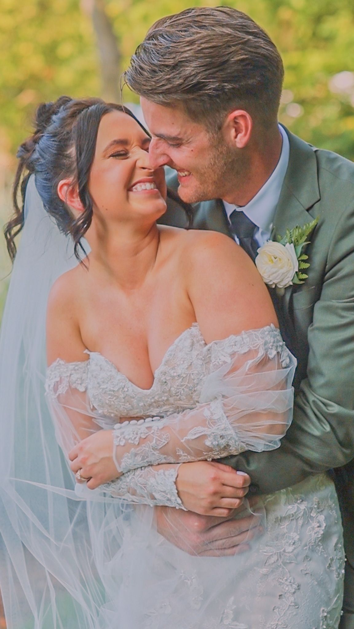 A romantic wedding photo of a couple embracing outdoors, with the bride in a lace dress and veil, and groom in a sage green suit.