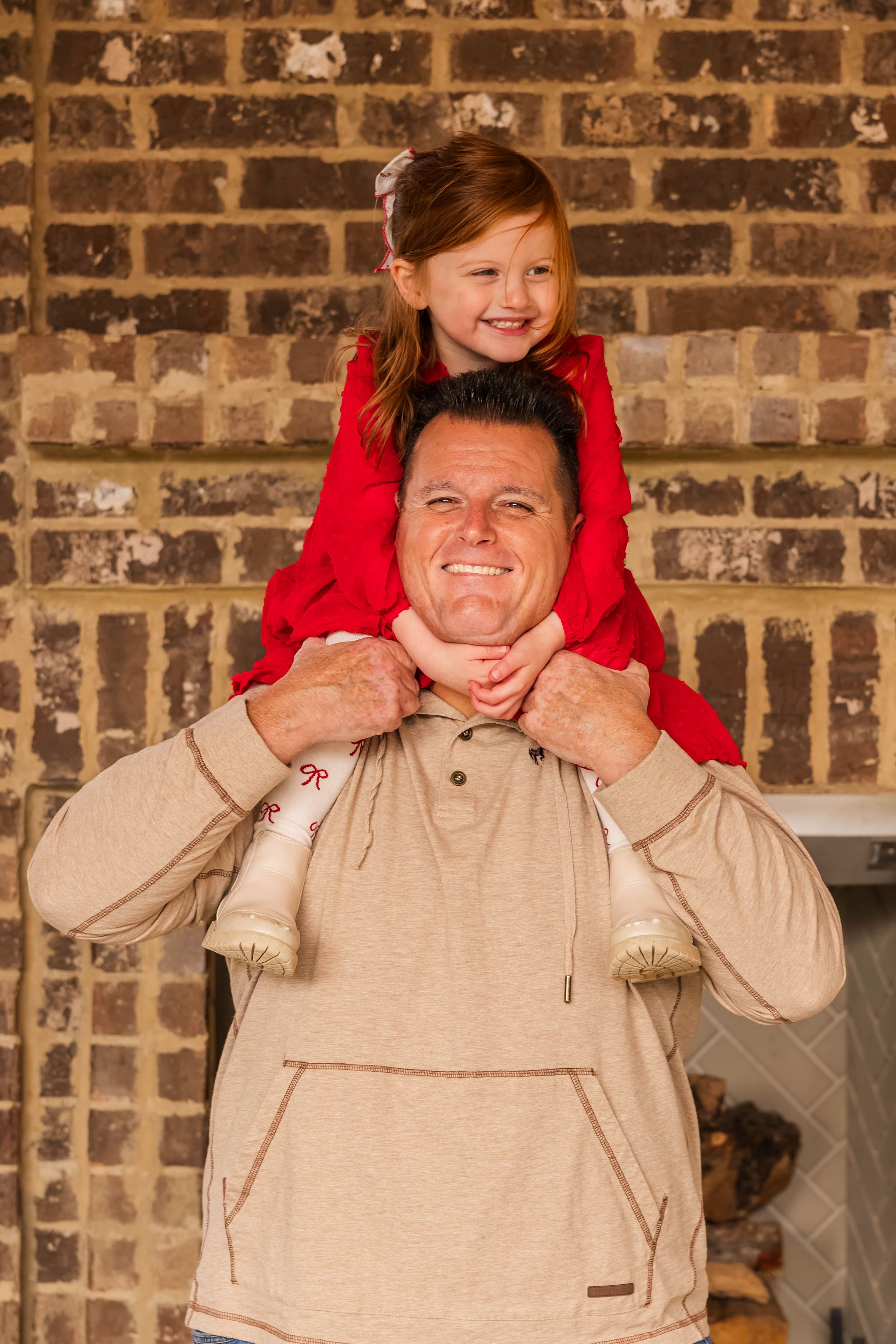 Child in red sits on adult's shoulders against brick wall background.