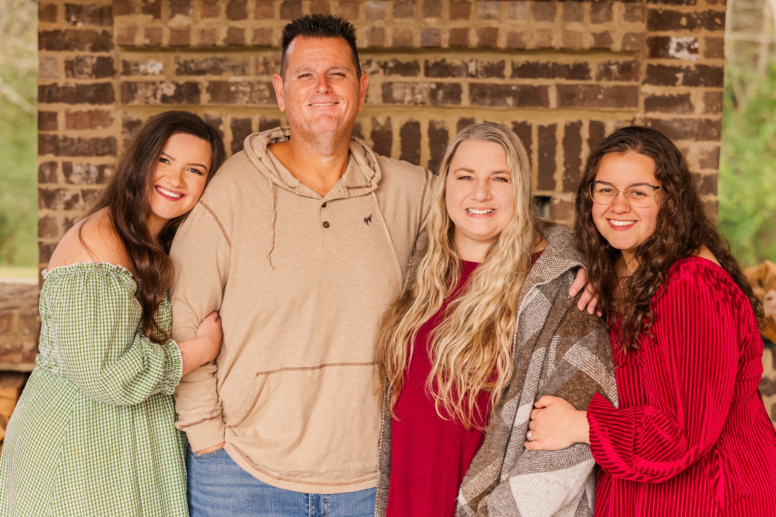 A family group poses together against a brick wall wearing casual sweaters in green, beige and red tones.