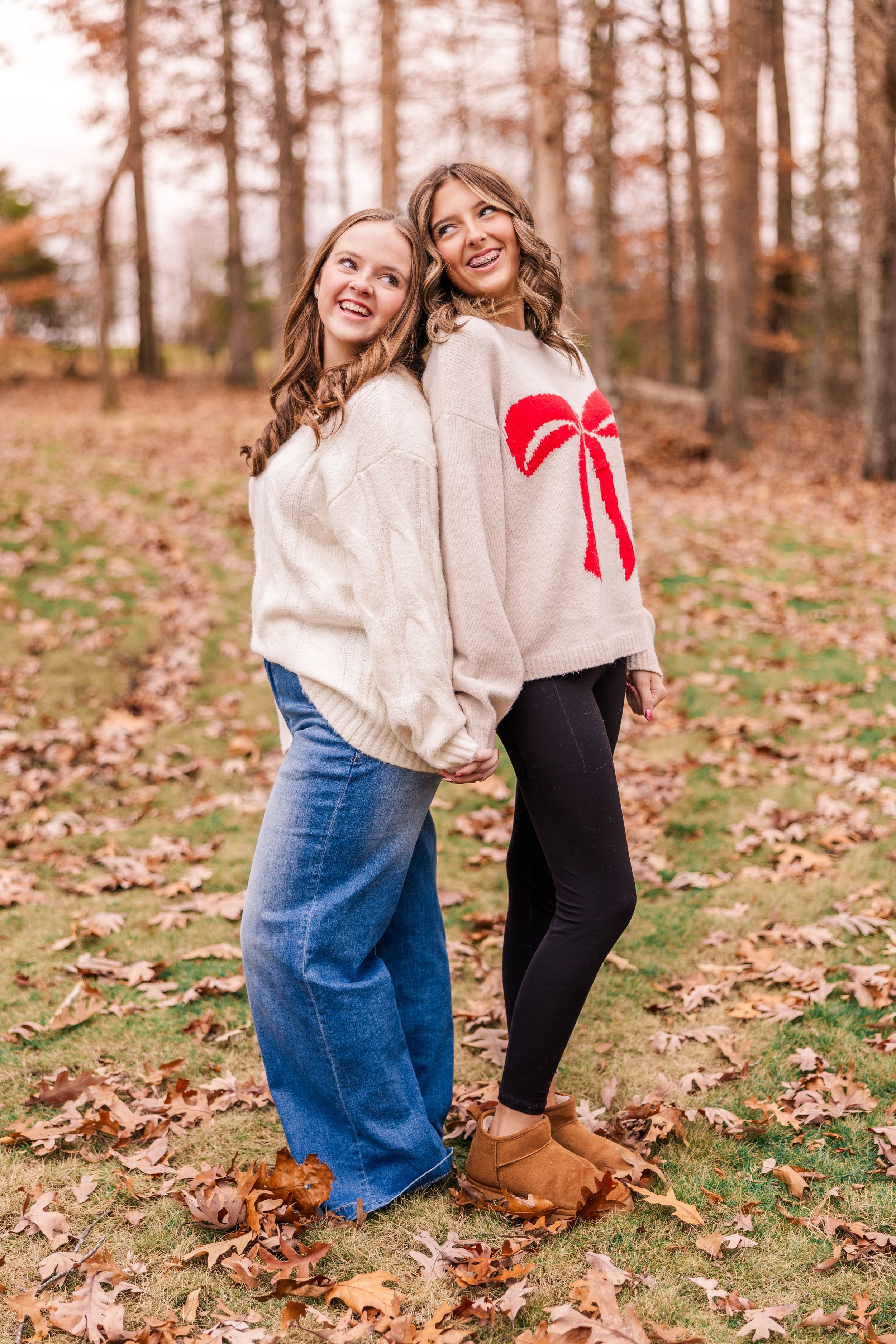 Two friends in cozy sweaters share a laugh while standing on fallen autumn leaves in the woods.
