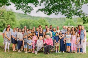 Large family group photo with multiple generations gathered outdoors under green trees.