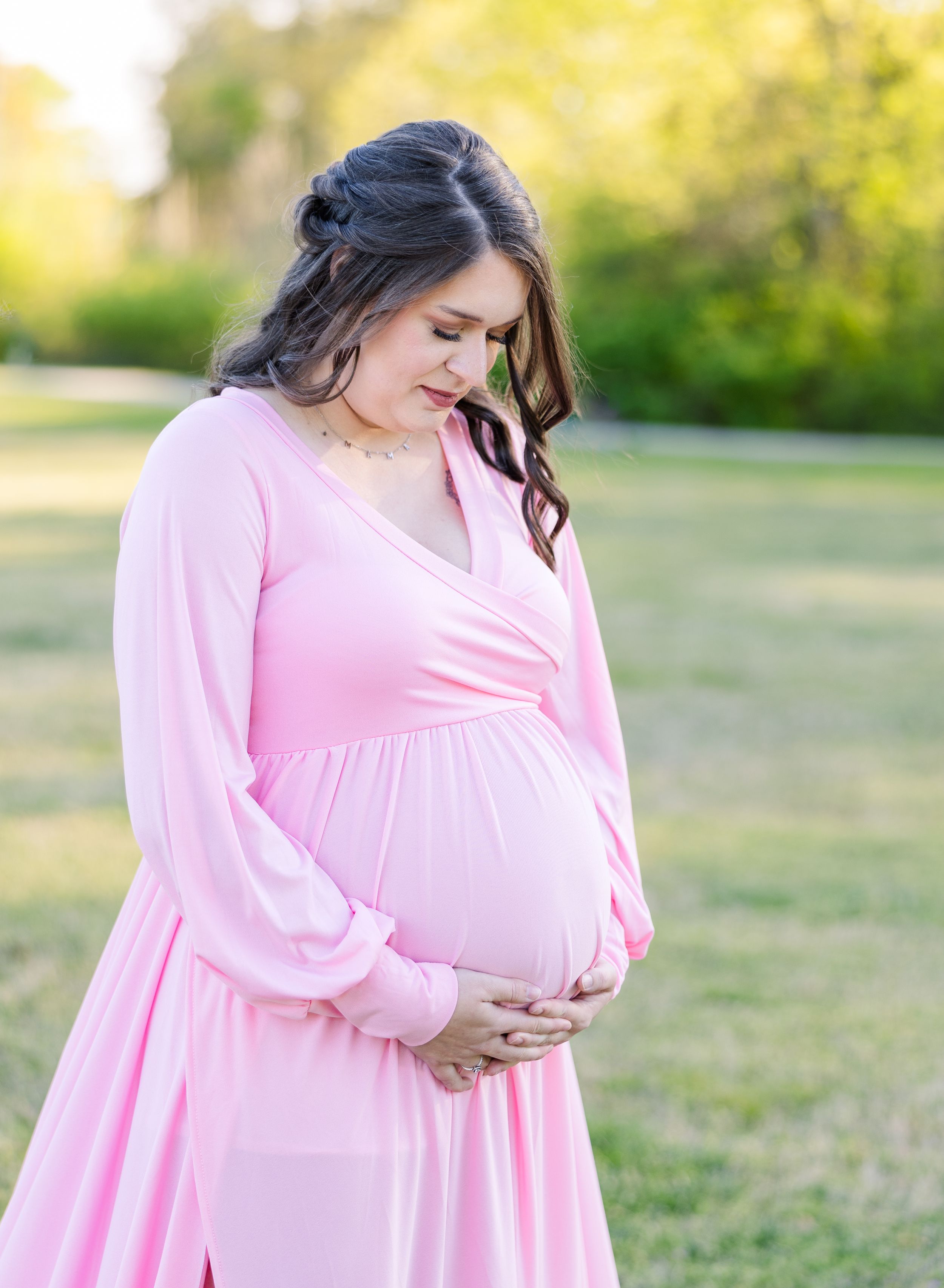 Maternity photo showing expecting mother in flowing pink dress at sunset in a grassy field.