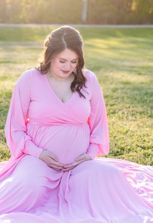 Pregnant woman in pink maxi dress sitting gracefully on grass during golden hour.