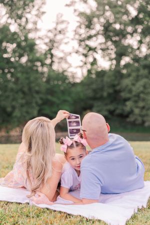 A family lies on a white blanket in the grass looking at photos together.